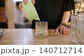 Matcha latte making. Barista work. Man pouring green liquid in glass adding milk from metal pitcher on wooden cafeteria bar counter. 140712714