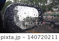 Slightly shaky handheld view from inside a car during a storm. Heavy raindrops stream down the window glass framing a side wing mirror as vehicles drive past outside. 140720211