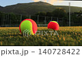 A womans hand throws orange tennis balls onto the green grass of a stadium at sunset, with beautiful mountains and a sports stadium in the background. 140728422