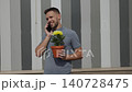 Caucasian man talks on phone while holding flower in simple terracotta pot against striped wall outdoors. Combining work life balance and family call by gift for mom, flower therapy for mental health. 140728475