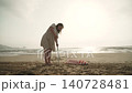 Caucasian woman independently installs the base of beach umbrella into sand on seashore. Person sticks umbrella pole on empty beach at sunrise. 140728481