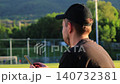 White man using smartphone by football field at sunset with athletes training in the background. Concept of phone addiction, digital distraction, social isolation, procrastination, modern screen 140732381