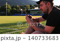 A white man sits relaxing and unwinding on the green grass of a football stadium at sunset, browsing his mobile phone. Concept of leisure time, summer evening, sports field, urban recreation, outdoor 140733683