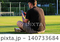 White man sitting on grass at a football stadium browsing smartphone while other players play football in the background at sunset. Concept of phone addiction, social isolation, social anxiety, sports 140733684