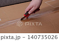 A hand using a utility knife to slice through adhesive tape on a cardboard shipping box, representing an order delivery arrival, unpacking process, and e-commerce shopping. Zoom 140735026