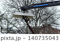 A hydraulic cherry picker lift with a metal basket extending upwards towards bare tree branches, depicting industrial height maintenance equipment in a quiet urban outdoor environment 140735043
