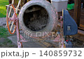 A concrete mixer mixes concrete in the backyard. Efficient mixing of concrete in drums. Close-up of the concrete mixer. Lawn and wooden scaffolding in the background. Uniform mixing process. Banner 140859732