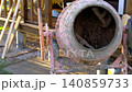A concrete mixer mixes concrete in the backyard. Efficient mixing of concrete in drums. Close-up of the concrete mixer. Lawn and wooden scaffolding in the background. Uniform mixing process. Banner 140859733