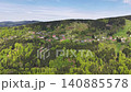 Aerial view village nestled in dense forest with red roofs, clearings, winding paths, mixed canopy and distant 140885578