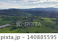 Aerial village nestled in valley with patchwork fields, rooftops and winding paths under soft clouds, drone 140885595