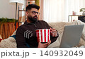 Indian man watching movie on laptop and eating popcorn at home, smiling and enjoying leisure time 140932049