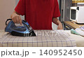 Hands guide a steam iron over a checked dishcloth on an ironing board in an indoor kitchen. 140952450
