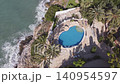 An aerial view captures a beachside property with a large pool. Waves crash against the rocks near the shore. Palm trees surround the area. The sun shines brightly above. 140954597