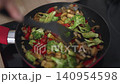 Chopping and stirring potatoes and broccoli with red bell peppers in a pan. This takes place in a kitchen during evening hours, showing colorful ingredients being cooked together. 140954598