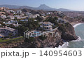 The camera shows various homes along a steep coastline in Spain. Many houses have pools and green areas. The ocean waves crash near the cliffs under the clear sky. 140954603