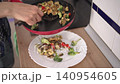 A person prepares a stir fry by cooking meat and vegetables together in a pan. They serve the dish on a plate and use a spatula to mix the ingredients well. 140954605
