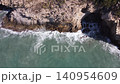 Waves hit the rocks at the coast. The sun shines on the clear water. This takes place during summer. The camera shows the shore from above. 140954609