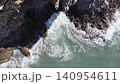 Waves hit the rocky shore as the sun shines down. The camera captures the movement of water and the textures of the rocks. The view shows the coast from above. 140954611