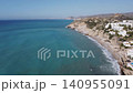 A drone captures a wide view of a beach with clear blue water and gentle waves. Sunlight shines on the shore as the camera moves over the coastline and the hills. 140955091