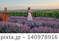A beautiful pregnant woman walking a blooming lavender field wearing a light dress and straw hat, she looks out over a green cornfield at sunset. Summer evening rural landscape 140978916