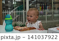 A baby girl with short blond hair takes a bite out huge pizza on the table, chews, picks corn with her fingers puts mouth. She chews appetizingly, squints sun, looks around. 141037922