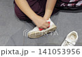 A person cleans a white shoe while sitting cross legged on a gray tiled floor. 141053956