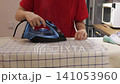 A steam iron moves over a checked kitchen towel while a person steadies the fabric on an ironing board. 141053960