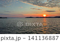A calm, warm sunset over the Aegean Sea in Gulluk, Turkey. The sun hangs low in a hazy orange sky, casting a bright golden light path across the gently rippling water toward distant dark hills. High 141136887