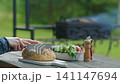 Close-up view of hands slicing fresh artisan bread in backyard with barbecue in background 141147694