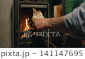 Close-up view of hand places firewood into log burner during evening at cozy wooden cabin. Flames rise inside wood stove creating warmth. Simple task for heating space. 141147695