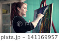 Beautiful redheaded Latina painting a picture in her studio. A very colorful painting, standing next to the window on a sunny day, wearing a denim overall. independent woman with attitude and beauty  141169957