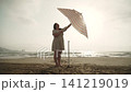 Caucasian woman in dress opens beach umbrella on sandy seashore on empty beach and sets up. Important UV protection during rest and swimming with tanning. Aesthetic shot of carefree vacation routine. 141219019