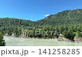 Wide video of a small red helicopter flying in the distance against a steep mountain covered with dense green pine forest, with a fast-flowing turquoise river in the foreground under a clear blue sky 141258285