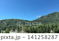 Small red helicopter flying high in the vast blue sky above a range of steep mountains densely covered with green coniferous trees 141258287