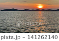 A calm, warm sunset over the Aegean Sea in Gulluk, Turkey. The sun hangs low in a hazy orange sky, casting a bright golden light path across the gently rippling water toward distant dark hills. High 141262140