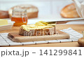 Soft butter spreading with a knife on a slice of fresh bread on wooden board, rustic kitchen atmosphere with loaf and honey jar in background, warm natural light, simple homemade breakfast concept 141299841