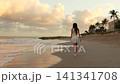 Woman strolling along a palm lined beach with beachfront buildings at sunset 141341708
