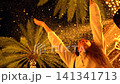 Woman raising arms beneath illuminated palm trees with falling snow 141341713