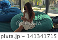 Woman relaxing on a bean bag chair using her smartphone by large windows 141341715