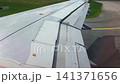 Aircraft wing with flaps in motion during landing approach, showcasing aerodynamic surfaces and surrounding green fields and taxiway markings on a sunny day 141371656