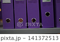 Upright purple binders on office shelf with one binder slightly pulled forward exposing narrow gap, circular finger holes, cardboard edges, neat storage, muted light, orderly side view 141372513