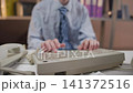 Office worker typing on beige keyboard while holding vintage telephone receiver beside bookshelves in focused desk-level workplace scene with business attire, muted colors, and indoor light 141372516