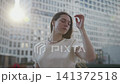 Confident woman turning and brushing hair in sunlit urban plaza beside tall office towers, soft backlight flare, gentle breeze, reflective modern city lifestyle 141372518