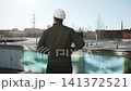 Rear view construction worker in white hard hat standing on rooftop edge overlooking industrial cityscape with pipes, towers, brick buildings, and clear daylight sky 141372521