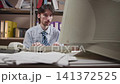Office worker typing at cluttered desk with oversized monitor in compact records room, showing focused data entry amid papers, folders, binder, printer, shelving and retro business workspace 141372525