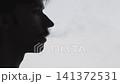 Silhouette profile of young man exhaling thin vapor plume against pale backlit background, tight sideview focusing on head and upper neck with negative space filled by dissipating cloud 141372531