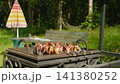 Meat skewers are prepared on grill. Blurred man sitting at wooden table in backyard and drinking beer on background 141380252