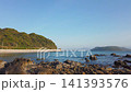 A scenic coastline with lush green hills and clear blue sky during a calm spring afternoon in April. Smooth footage captures a quiet bay with rocks in the shallow turquoise sea near Fukuoka, Japan.  141393576