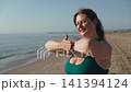 Caucasian woman brings together palms of outstretched hands in clasp on seashore in sunlight. Portrait of person doing exercises in morning on empty beach and enjoying inspiring moment with smile. 141394124