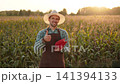 Portrait of happy farmer in hat and apron before cornhole game near farm field showing thumbs up. Confident agricultural worker during active break after work in cornfield. 141394133